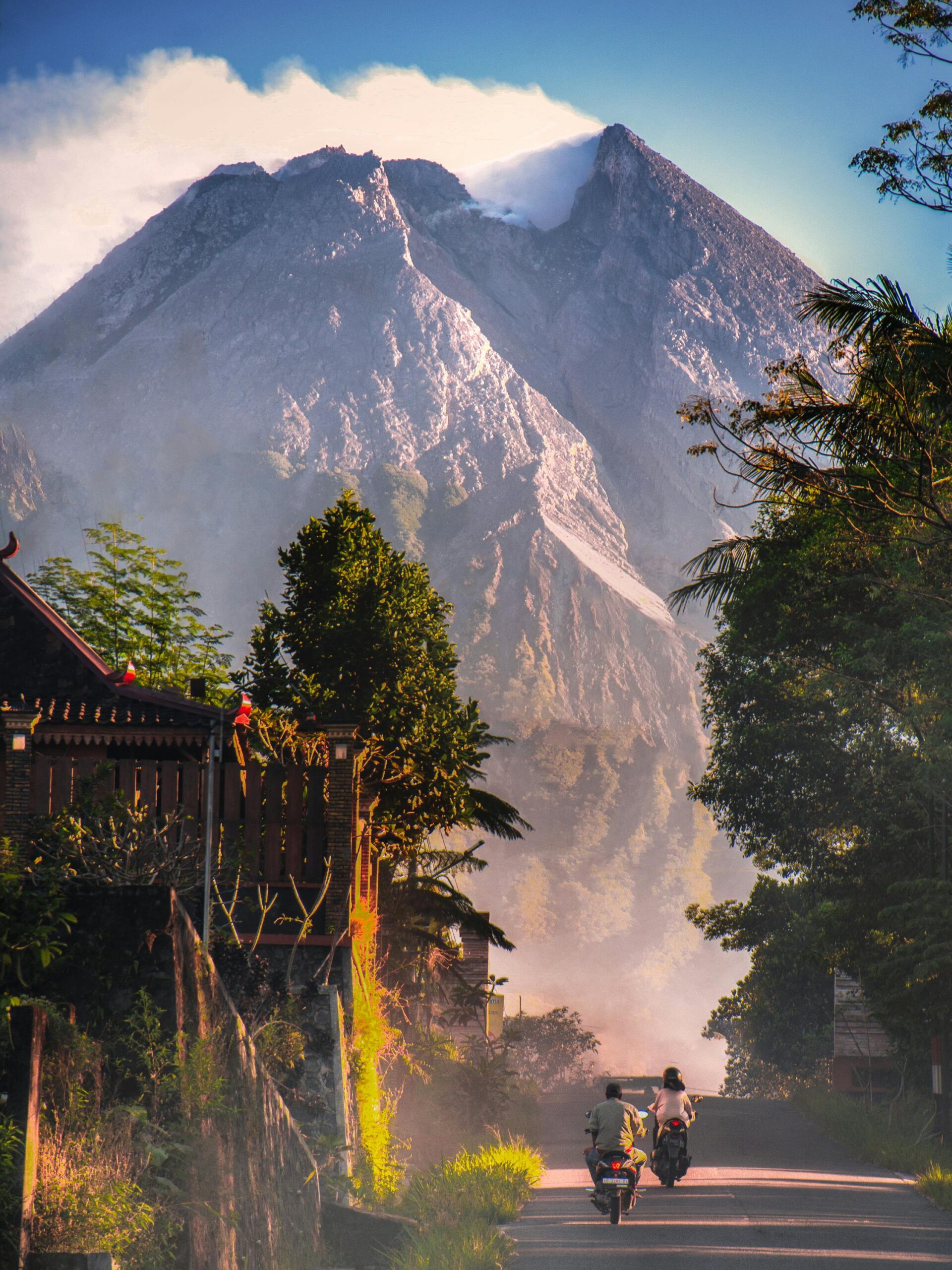 Home Motorcyclists ride towards a stunning volcano on a lush, tree-lined road under a clear sky.